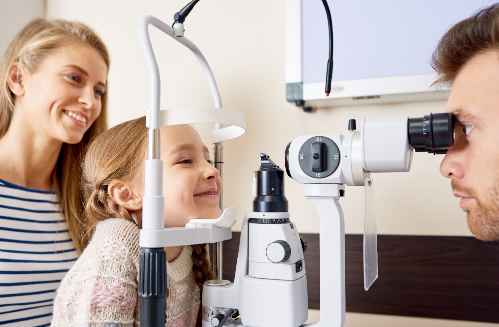 A smiling mother and daughter sit together as an optometrist gives the daughter an eye exam
