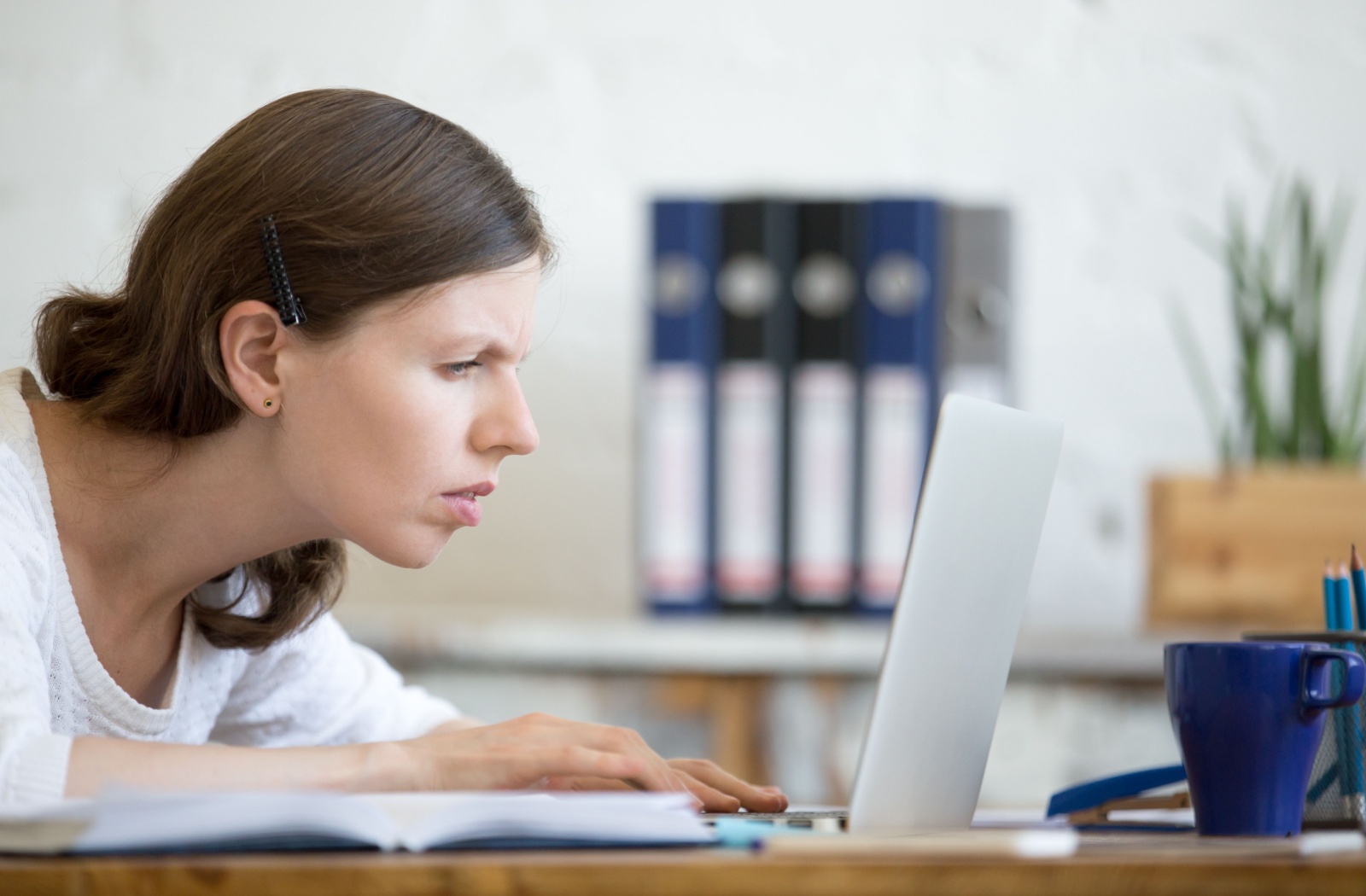 A young businessperson sits in a well-lit office, squinting closely at their laptop.
