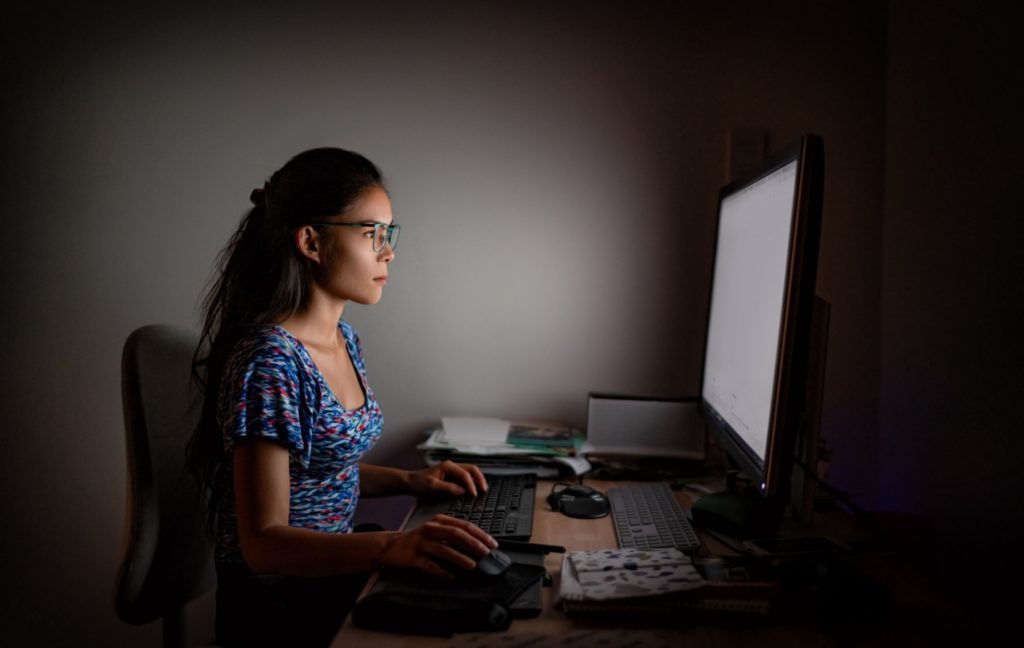 A person wearing glasses is lit by their computer screen as they work at their desk in a dark office.