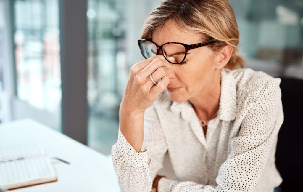 An older adult lifts their glasses and pinches their eyes to clear blurry vision caused by dry eyes.