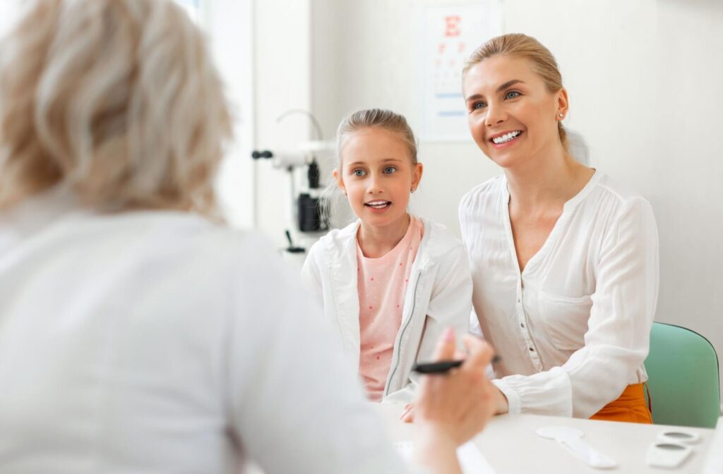 A smiling mother and daughter talking with an optometrist during an eye care consultation, sitting together in a bright clinic with vision testing equipment in the background.