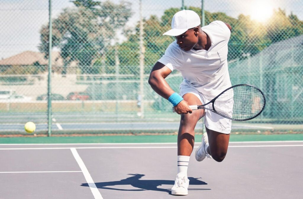 Tennis player in white athletic gear and cap lunging forward on an outdoor court to hit a tennis ball, captured mid-action under bright sunlight.